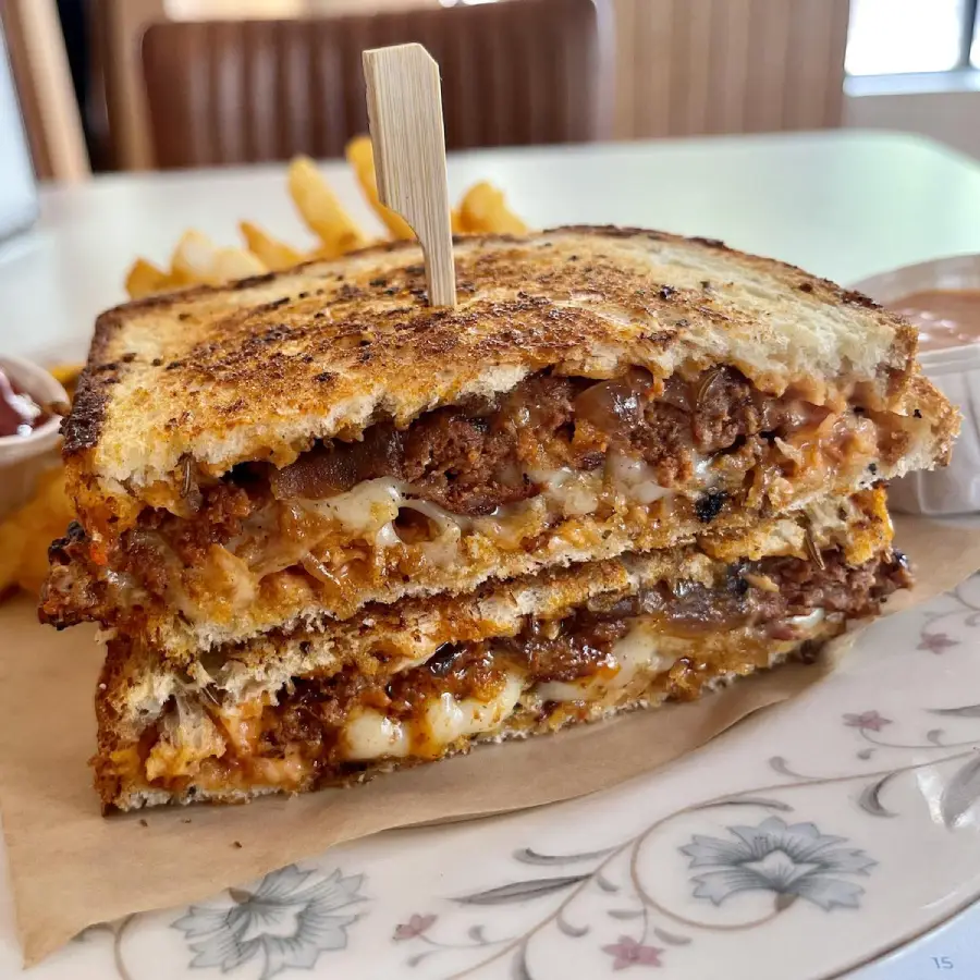 A close-up of a toasted sandwich cut in half, showing melted cheese, meat, and sauce inside, held together by a wooden stick. French fries and a dipping sauce are visible in the background.