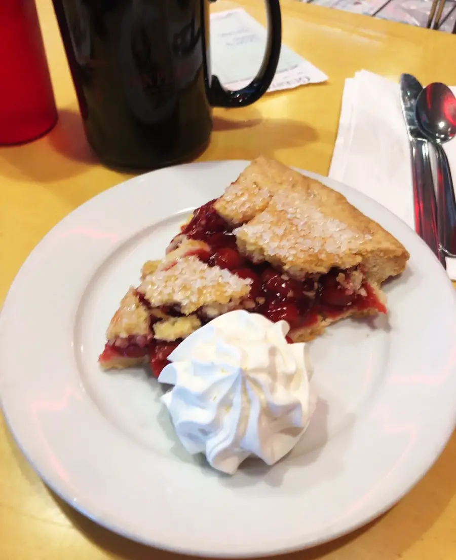 A slice of cherry pie with a crumbly crust is served on a white plate with a dollop of whipped cream, next to a mug, utensils, and napkins on a yellow table.