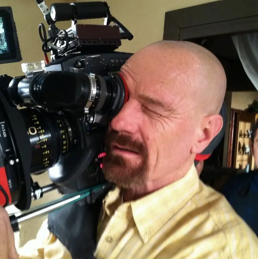 A bald man with a goatee, wearing a yellow shirt, looks through the viewfinder of a large professional film camera, appearing to operate it on a set indoors.