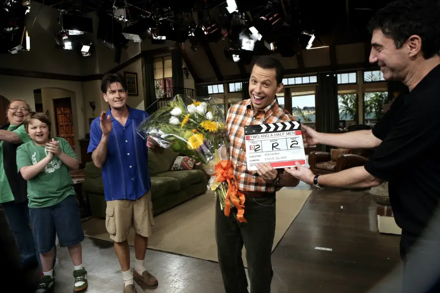Four people smile and applaud on a TV set. One man holds flowers and a clapperboard, while others cheer. Studio lights and cameras are visible above a living room set. The mood is celebratory and joyful.