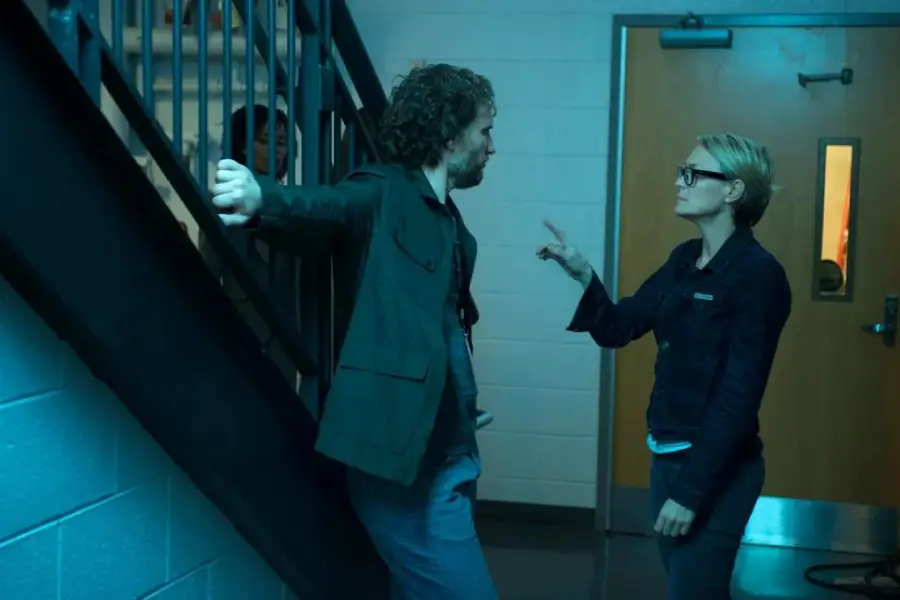 A man leans against a stair railing while a woman wearing glasses stands facing him, pointing her finger as they have a serious conversation in a hallway lit with blue light.
