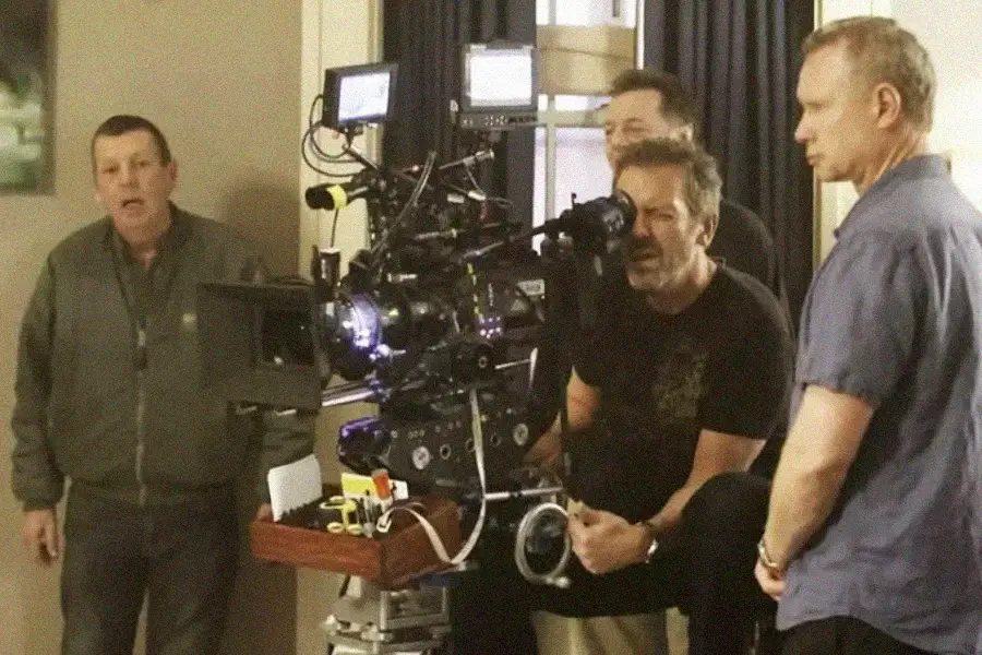 A film crew operates a large camera setup indoors; three men focus on the camera, while another stands to the side, observing the scene.
