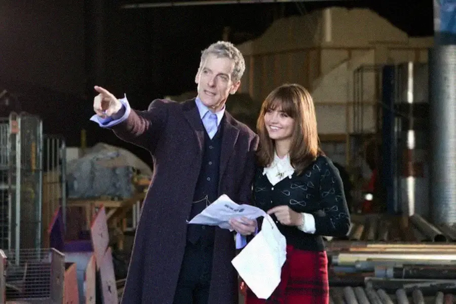 A man in a long dark coat points ahead while holding papers, standing next to a smiling woman with brown hair who also holds papers. They appear to be on a set, surrounded by industrial and construction materials.