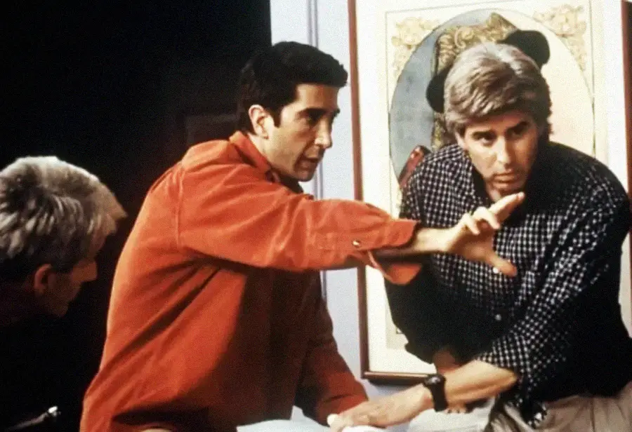 Three men are gathered around a table, one in an orange shirt gesturing with his hand as if explaining something to another man in a checkered shirt. A framed artwork hangs on the wall behind them.