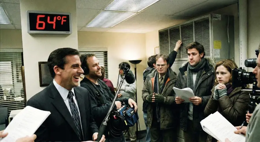 A man in a suit laughs while surrounded by people with scripts and film equipment in an office setting. A digital clock on the wall displays 64°F. Others appear to be preparing for a scene, holding papers and a microphone.