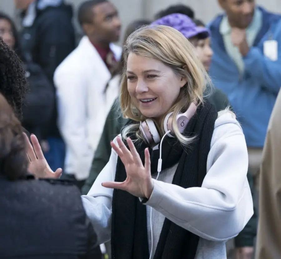A woman with blonde hair, wearing a light hoodie, scarf, and headphones around her neck, smiles and gestures with her hands while talking to someone outdoors among a group of people.