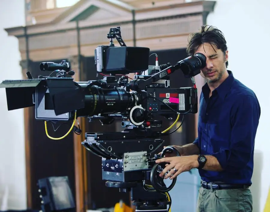 A man in a navy shirt operates a large professional film camera on set, looking through the camera's viewfinder and adjusting its controls, with filming equipment visible in the background.