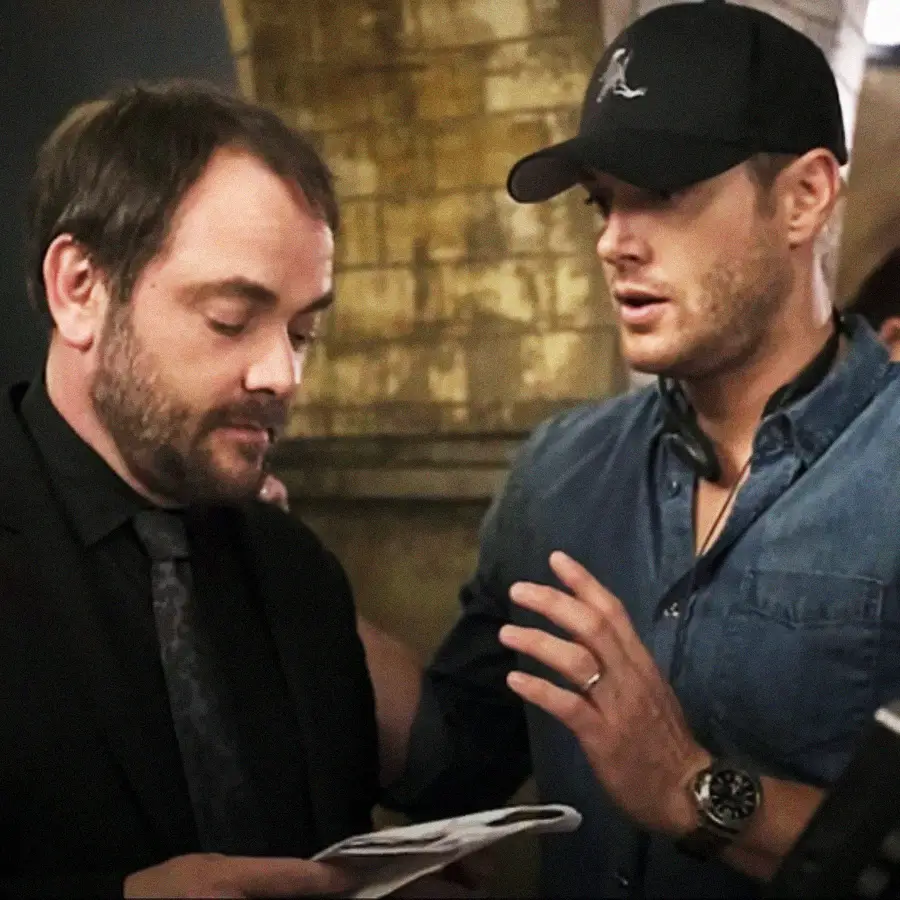 Two men stand close together indoors. One wears a black suit and looks down at papers, while the other, in a blue shirt and black cap, gestures with his hand as if explaining something. The background is blurred.