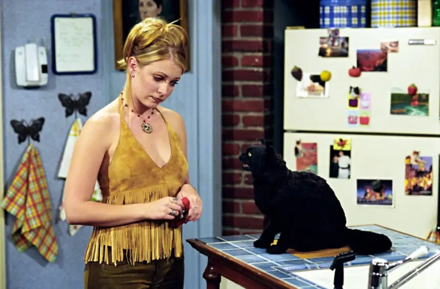 A young woman with blond hair in a ponytail and a fringed suede top stands by a kitchen counter, looking at a black cat sitting on the counter. A fridge with photos and magnets is in the background.
