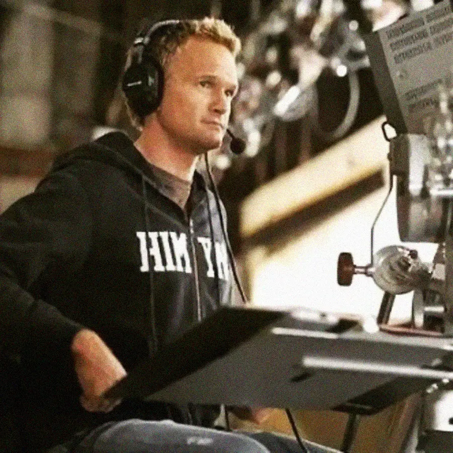 A person wearing headphones and a dark hoodie sits at a desk with a microphone and monitor, appearing focused, possibly working behind the scenes in a production environment.