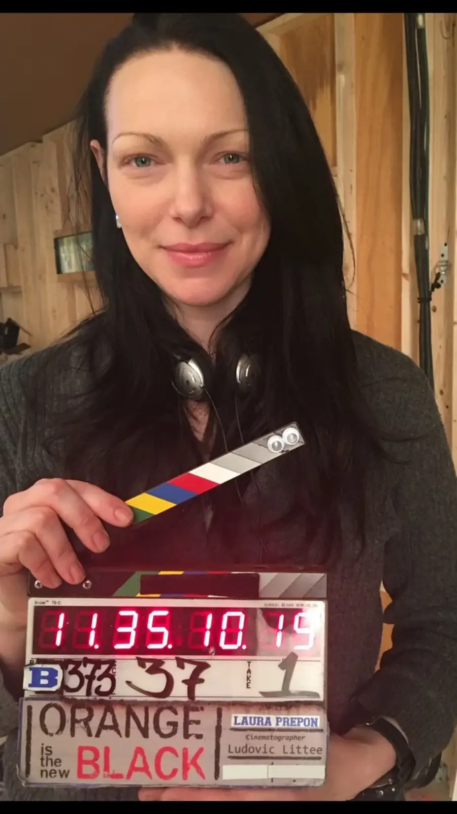 A woman with long dark hair and headphones around her neck smiles while holding a film clapperboard labeled “Orange is the New Black” in front of her. The background appears to be a wooden set.