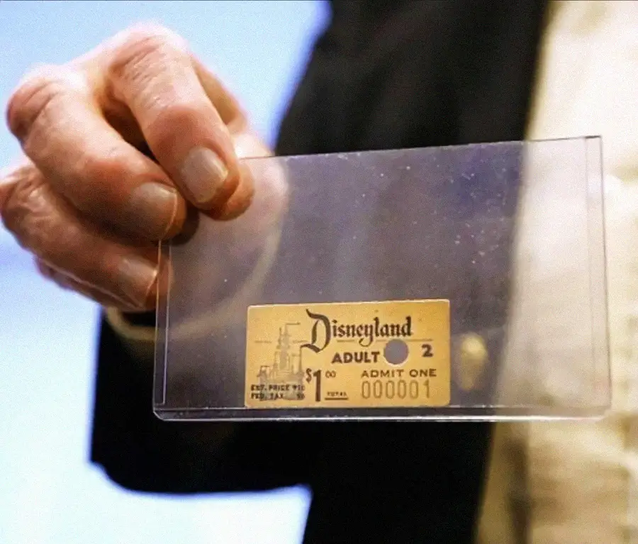 A person holds up a transparent case containing an old Disneyland admission ticket labeled "Adult 2, Admit One, 000001" with a small illustration of the park.