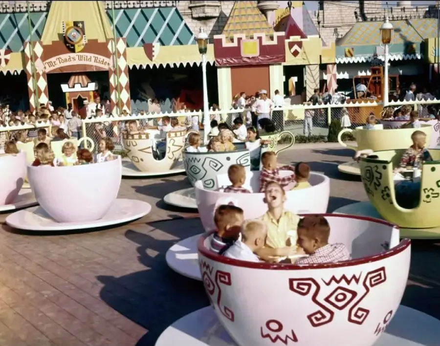 Children and adults ride spinning teacups at an amusement park attraction, surrounded by colorful, whimsical buildings and a crowd of onlookers. The atmosphere appears lively and cheerful.