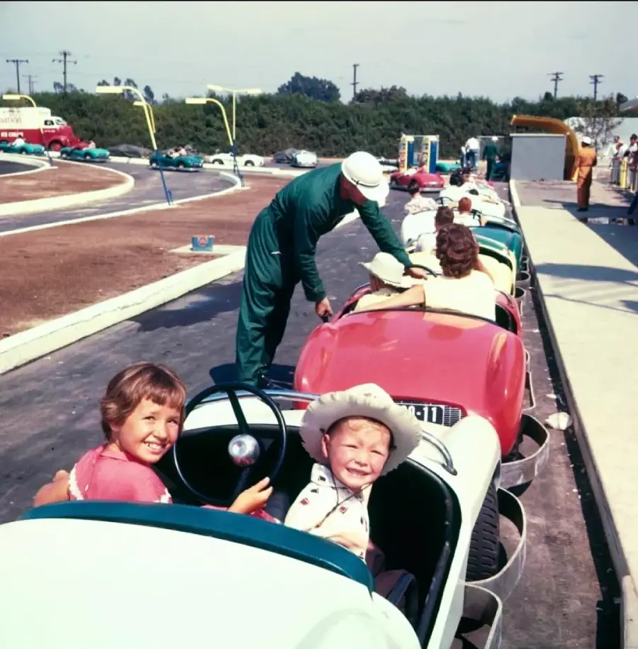 Children sit in small colorful cars on an amusement park ride, smiling at the camera. An adult attendant in green helps a rider. The track curves, and other people watch nearby. It's a sunny day with blue sky and trees in the background.