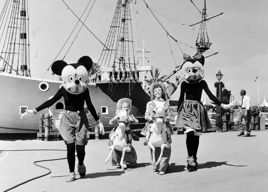 Four people walk side by side; two in large Mickey and Minnie Mouse costumes, and two children in cowboy outfits riding stick horses, with a large ship in the background. The scene appears to be at a theme park.