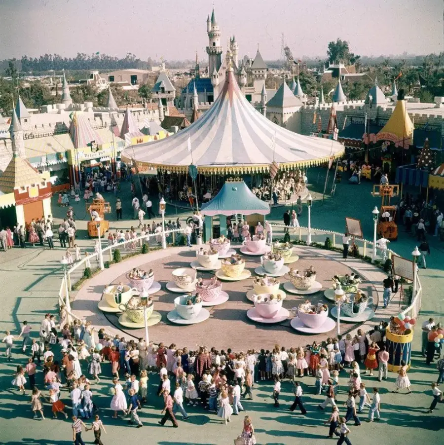 A vintage photo of Disneyland shows crowds around the Mad Tea Party teacup ride, with pastel teacups spinning under a striped canopy. The park is busy, with Fantasyland buildings and Sleeping Beauty Castle in the background.
