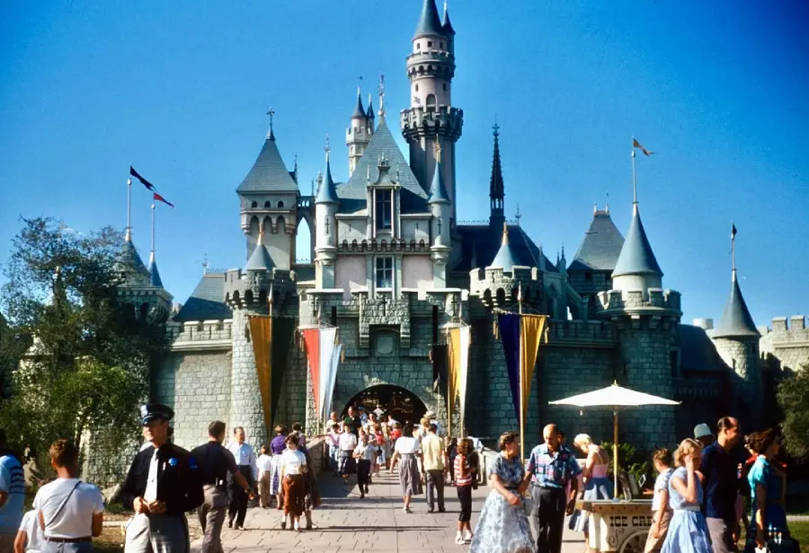 Visitors walk in front of a large fairytale castle with blue spires at Disneyland on a sunny day, with colorful banners, trees, and a clear blue sky in the background.