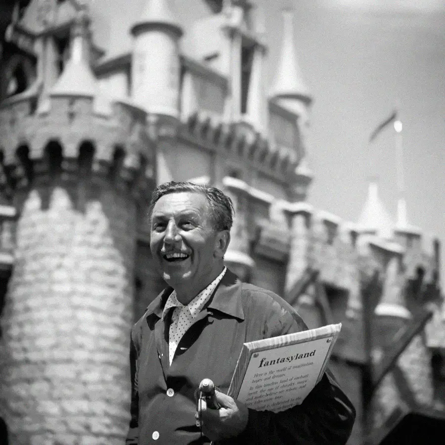 A man smiling and holding papers in front of a castle at an amusement park. The castle has stone walls and tall turrets, and the man appears cheerful and excited.