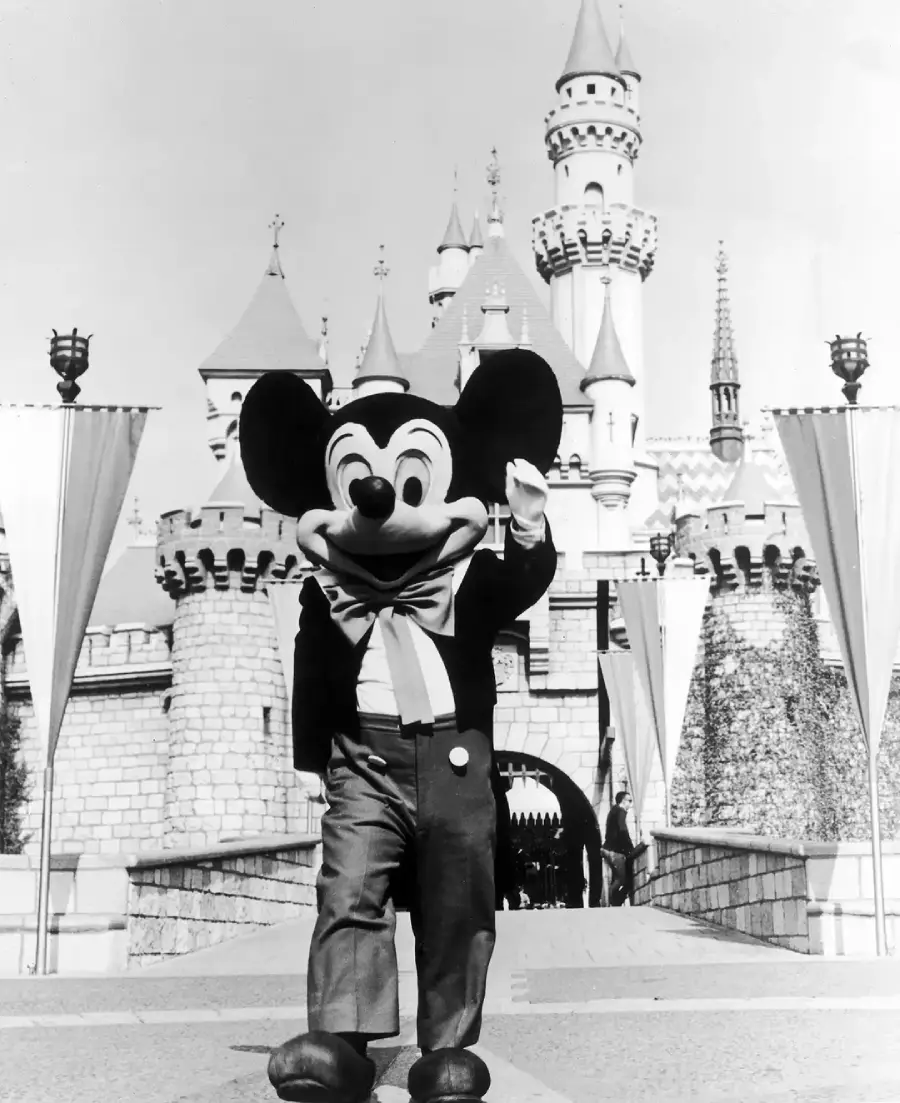 A person in a classic Mickey Mouse costume stands in front of Sleeping Beauty Castle at Disneyland, waving with one hand. The photo is black and white and features castle turrets and flags in the background.