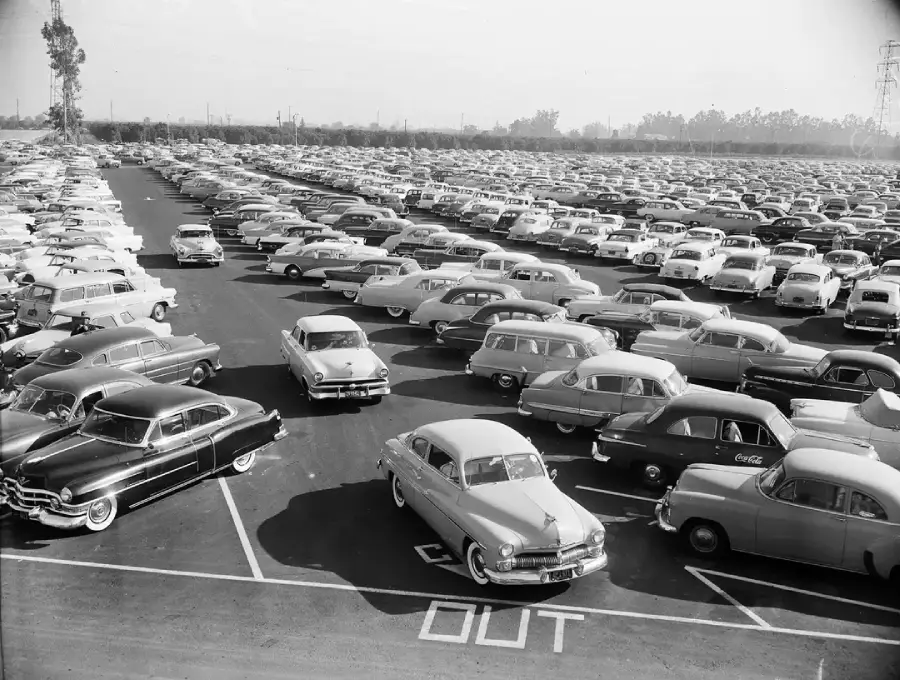 A large, crowded parking lot filled with classic 1950s cars, seen in black and white. Cars are parked in neat rows, with two cars near a painted "OUT" sign in the foreground.
