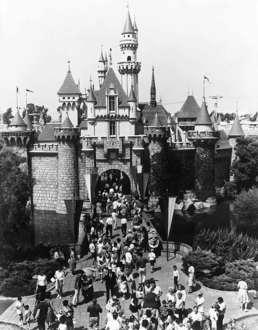 Black-and-white photo of a large crowd walking toward Sleeping Beauty Castle at Disneyland, with towers, turrets, and flags visible, surrounded by trees and landscaping.