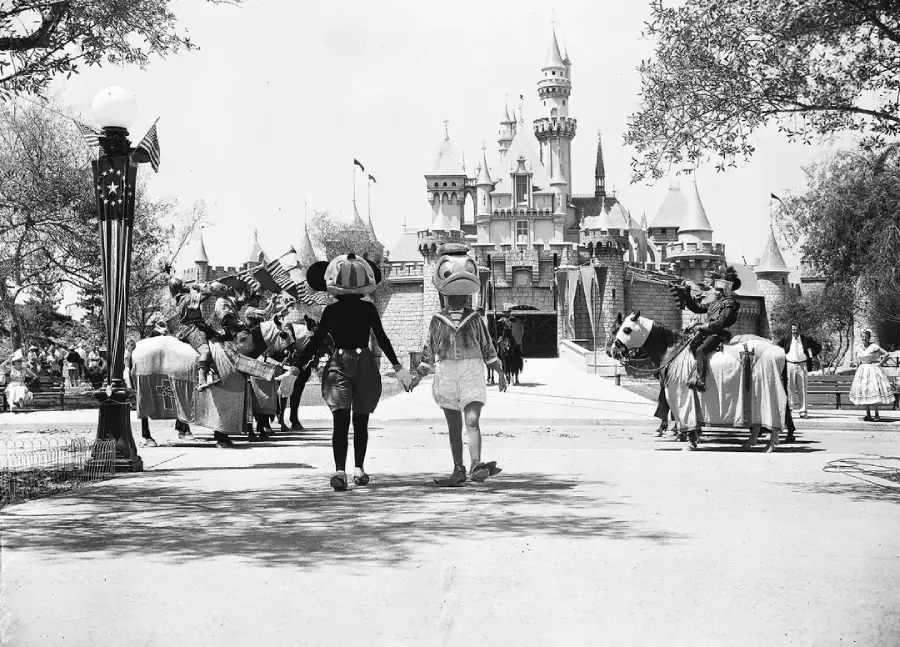 Two people in cartoon character costumes walk hand in hand toward Sleeping Beauty Castle at Disneyland, with other costumed characters and visitors around on a sunny day. The scene is in black and white.