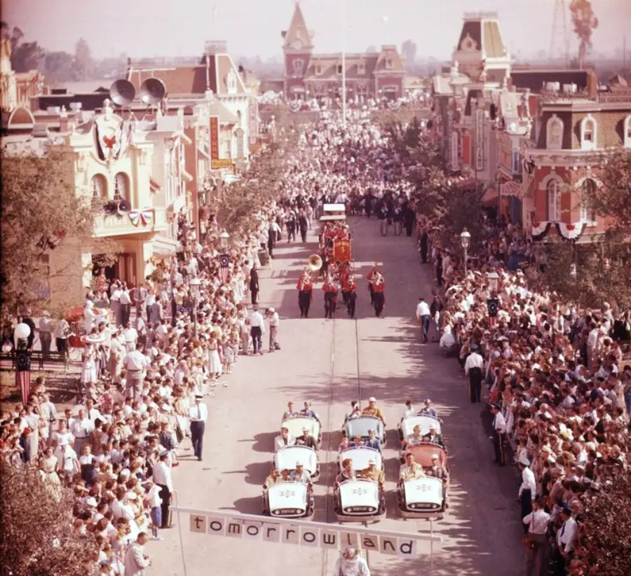 A vintage parade at Disneyland shows crowds lining Main Street, classic cars with police escorts, a marching band, and a banner reading “Tomorrowland,” with park buildings and Sleeping Beauty Castle in the background.
