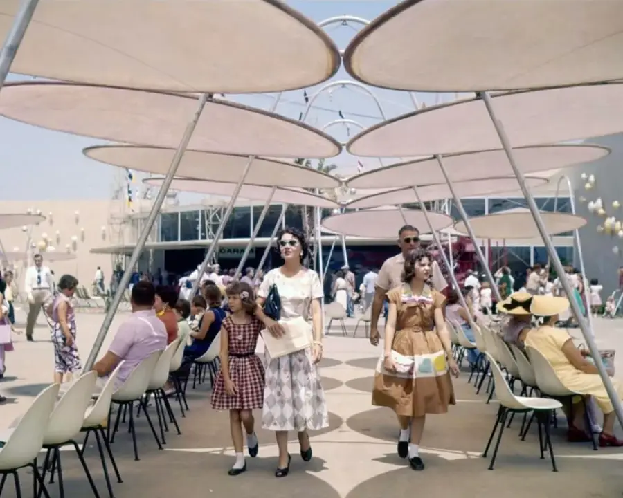People walk and sit under large, round sunshades at an outdoor event. A family dressed in vintage 1960s clothing walks through the center while others relax on white chairs. The scene is lively and sunny.