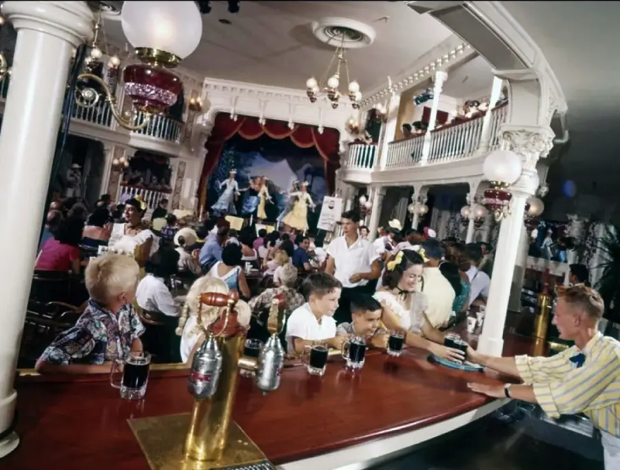 A lively, vintage-style saloon filled with people sitting at tables and at the bar, watching performers in colorful costumes dancing and singing on a stage in the background.