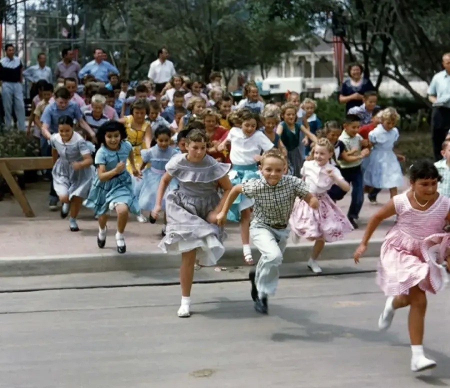 A large group of excited children in colorful clothes run toward the camera, while adults watch from the background in an outdoor park setting. The children appear cheerful and energetic.