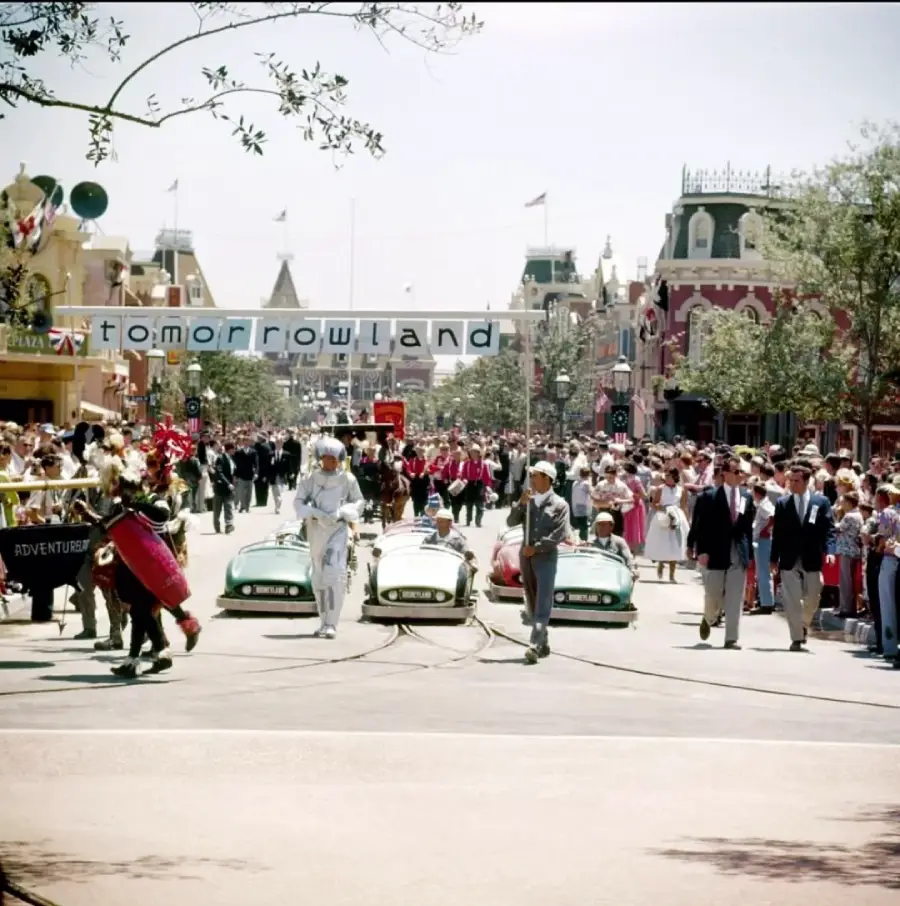 A vintage parade at Disneyland features people in futuristic costumes driving small cars under a "Tomorrowland" banner, with crowds of spectators and Main Street buildings in the background.