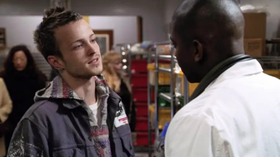 A young man in a gray jacket faces a man in a white coat in what appears to be a storage or laboratory setting, with shelves and people in the background.