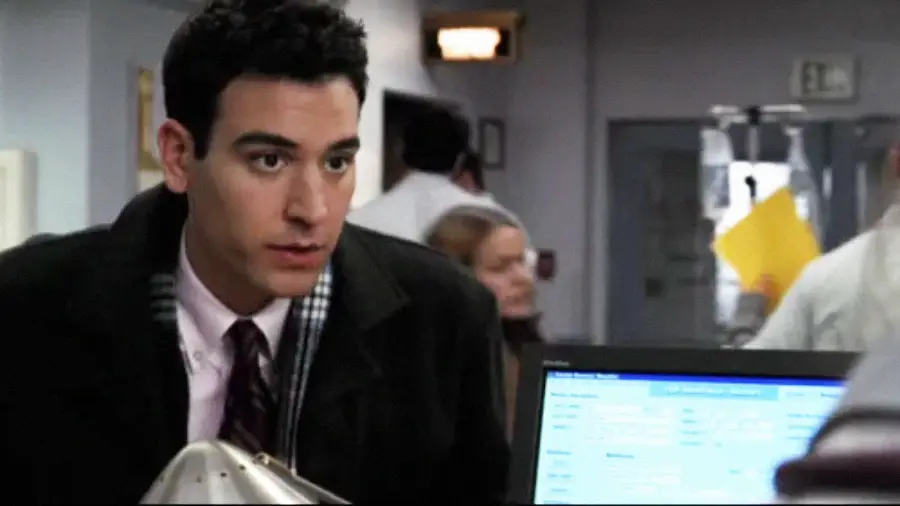 A man with dark hair in a suit and coat leans on a counter in what appears to be a hospital or clinic, with blurred staff and equipment in the background. A computer screen is visible in the foreground.