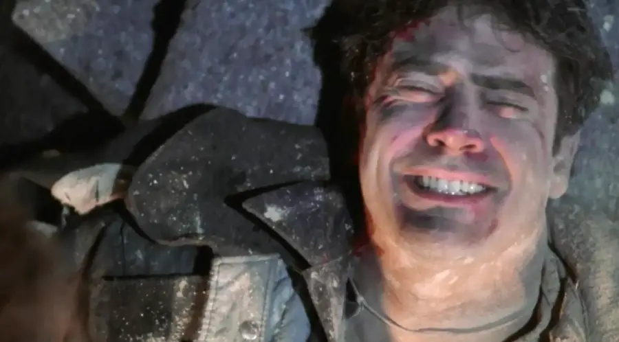 A man with dark hair lies on a rough surface, his face contorted in pain. He is covered in dust and debris, wearing a dark jacket, and appears to be injured.