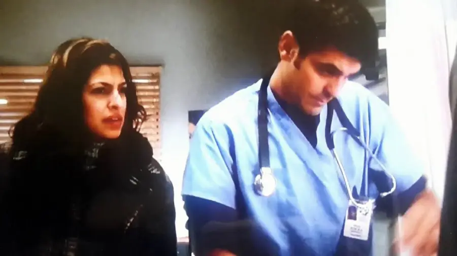 A woman with wavy hair stands next to a man wearing blue medical scrubs, a stethoscope, and an ID badge. The man appears to be performing a medical task while the woman looks on with concern.