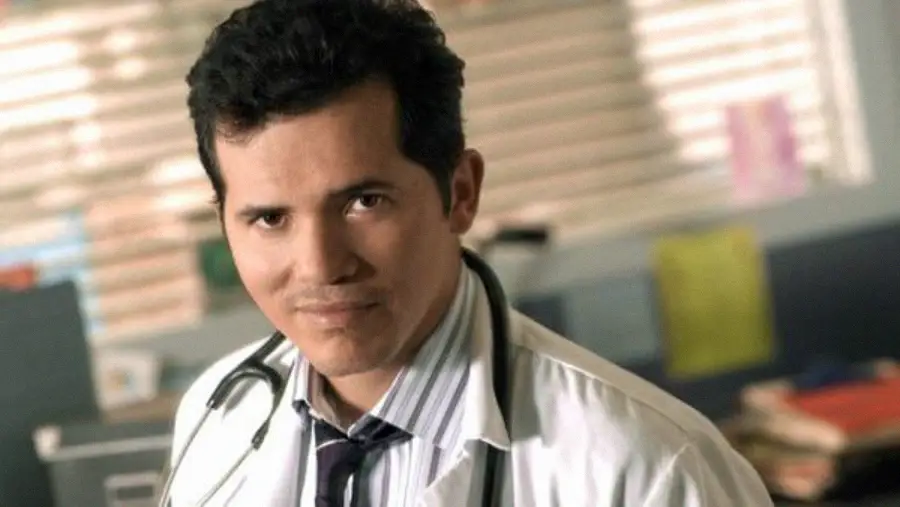 A man wearing a white doctor's coat and a stethoscope around his neck stands in an office with blinds and colorful folders in the background. He is looking at the camera and smiling slightly.