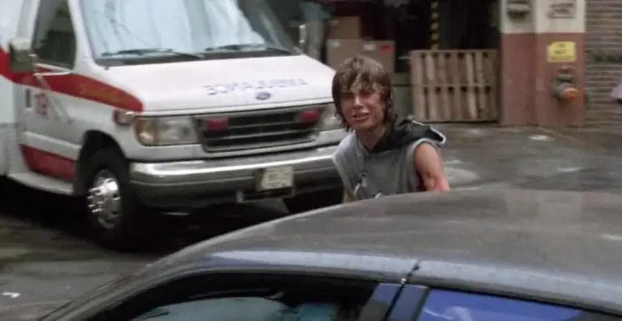 A young person with messy hair and glasses stands outdoors near a gray car, looking toward the camera. An ambulance is parked in the background, along with a wooden pallet and some boxes.
