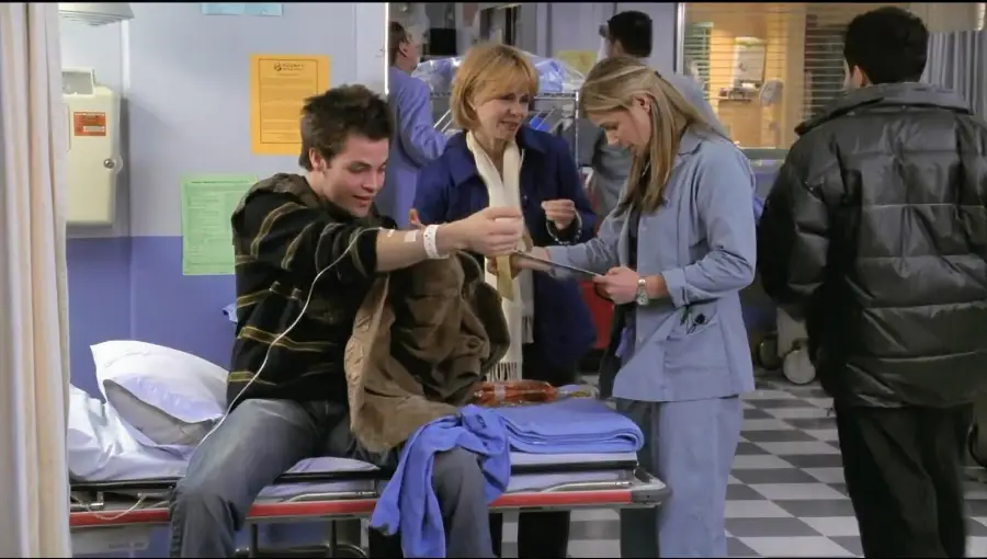 A man sits on a hospital bed with his sleeve rolled up as a nurse prepares his arm for treatment. A woman stands nearby, and other people are visible in the busy hospital hallway.