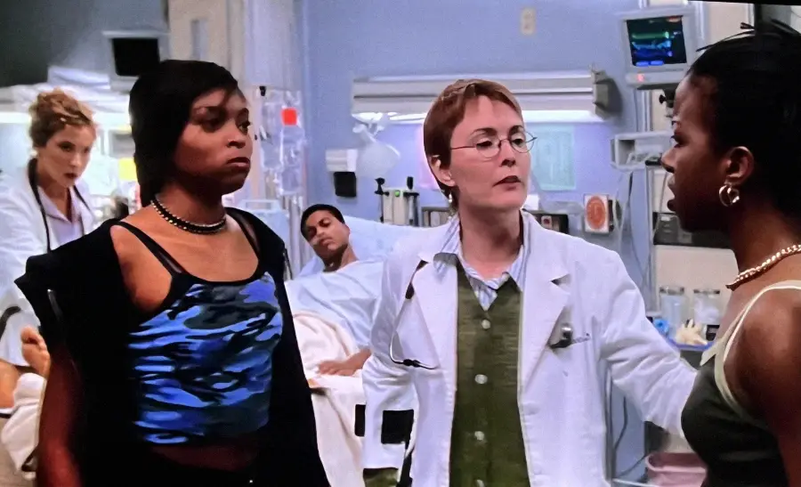 Two women stand facing each other in a hospital room, one wearing a blue tank top, the other a gold necklace. A doctor in a white coat stands between them, with a patient lying in a bed in the background.