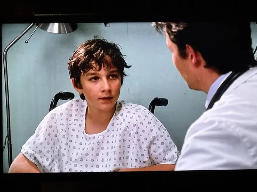 A teenage boy wearing a hospital gown sits in a wheelchair, facing a man in a white coat who appears to be a doctor, in a hospital room with gray walls and a lamp in the background.