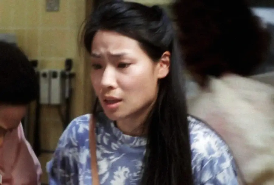 A woman with long dark hair wearing a blue and white patterned shirt looks concerned or worried in an indoor setting, with other people and blurred backgrounds visible.
