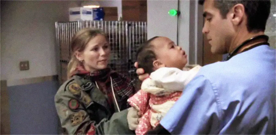 A man in medical scrubs holds a baby while a woman in a jacket and scarf looks on, standing in what appears to be a hospital or medical setting.