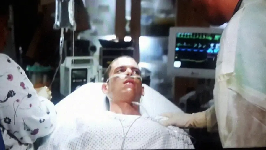 A young man lies in a hospital bed with a nasal cannula for oxygen and medical monitors in the background; two people in medical attire stand beside him, one holding his shoulder.