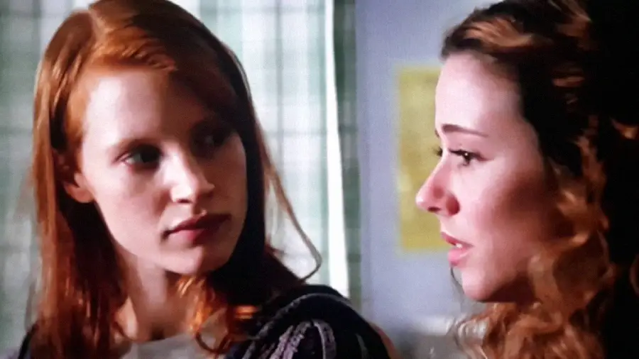 Two young women indoors facing each other; the woman on the left with straight red hair looks intently at the woman on the right, who has wavy brown hair and appears to be speaking or emotional.