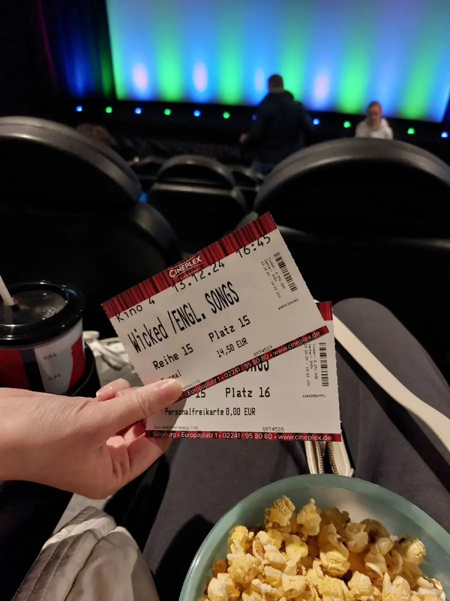 A hand holds two movie tickets inside a cinema, with seats, a drink, and a tub of popcorn visible in the foreground, and a colorful lit screen in the background.