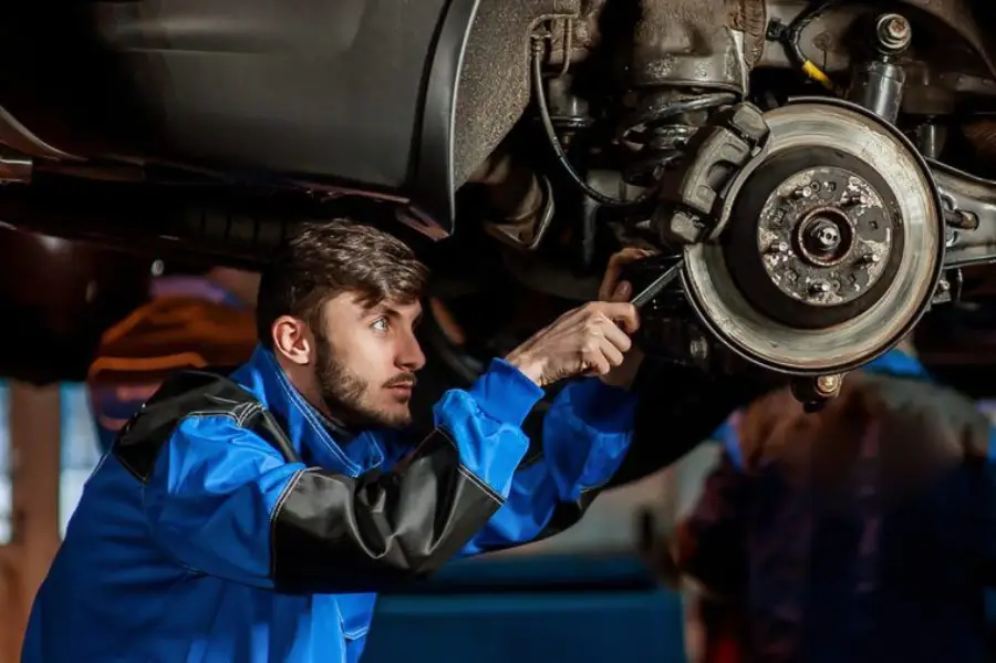 A mechanic in a blue and black uniform works underneath a raised vehicle, using a tool to inspect or repair the car’s brake system and suspension components.