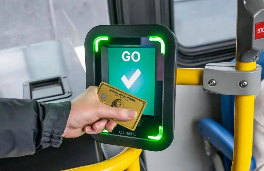 A person holds an American Express card to a bus fare payment terminal. The screen displays a green "GO" with a check mark, indicating successful payment. Bright green lights frame the screen.