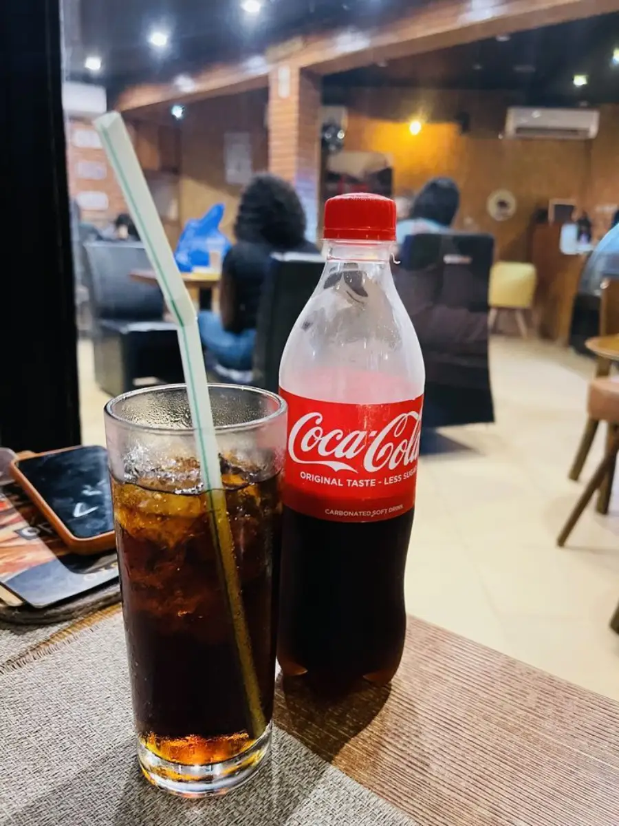 A glass of iced Coca-Cola with a straw sits next to a Coca-Cola bottle on a wooden table in a cozy café, with people seated and socializing in the blurred background.