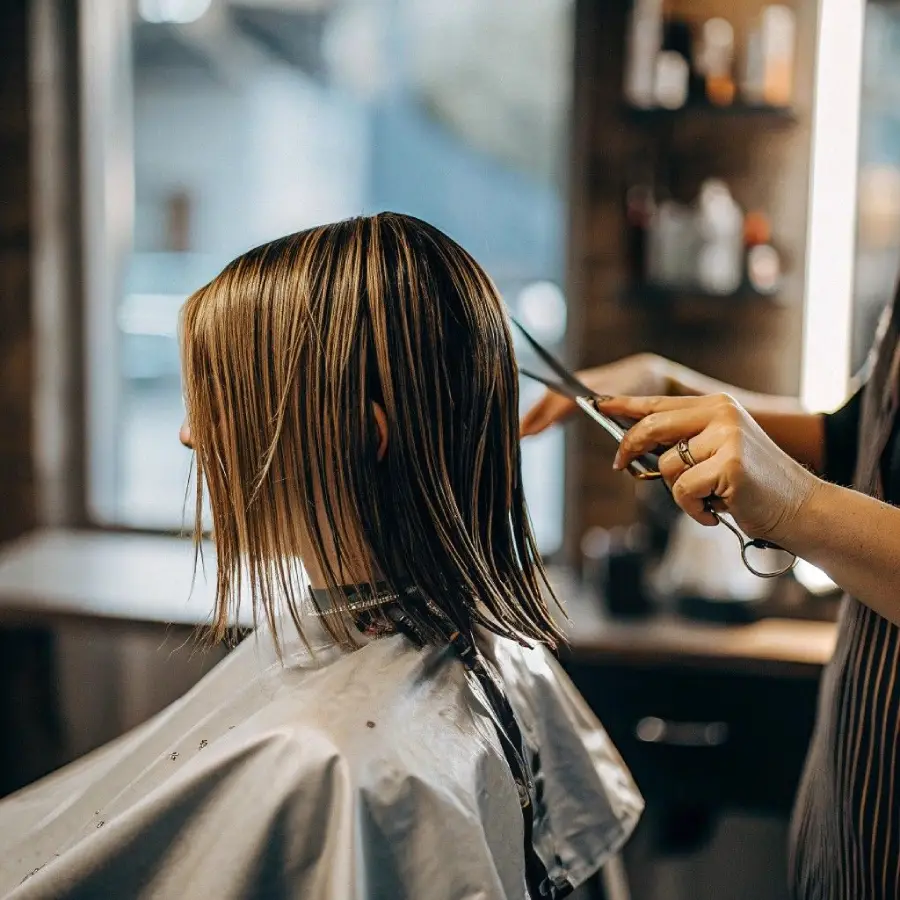 A person with straight, wet, shoulder-length hair sits in a salon chair while a stylist trims their hair with scissors. The stylist's hands are visible, and the salon interior is softly blurred in the background.