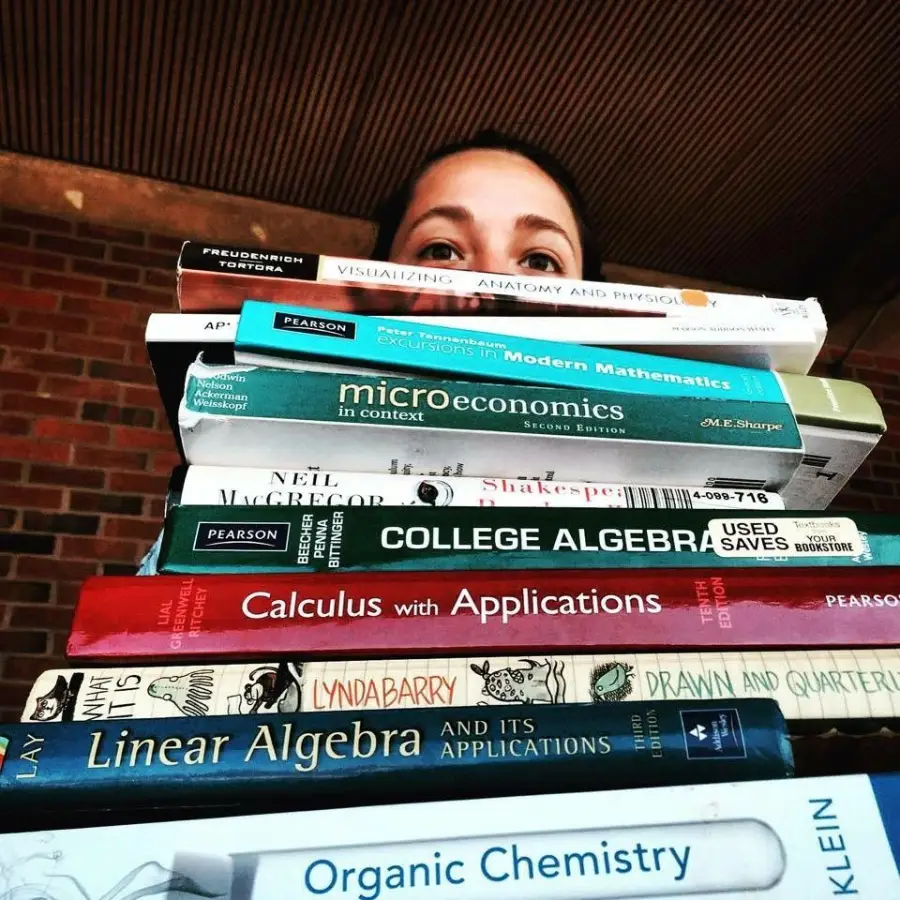 A person peers over a tall stack of textbooks, including subjects like anatomy, microeconomics, algebra, calculus, linear algebra, and organic chemistry, suggesting a heavy academic workload.